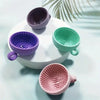 A collection of silicone makeup brush cleaners in purple, green, and pink sitting on a white surface.