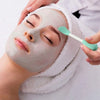 Hands apply a white silicone facial mask to a person's face under a towel.