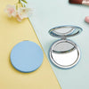 A light blue portable magnifying makeup mirror is shown on a pastel colored surface next to a pink flower.