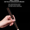 Une main tient un pinceau de correcteur avec une tête en demi-lune et un produit beige clair sur fond noir