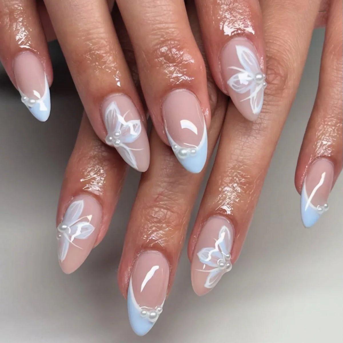 A close-up of a hand displaying 24-piece almond press-on nails featuring white and light blue floral and butterfly designs.