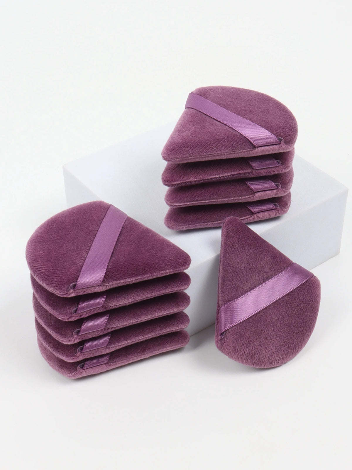 A stack of triangular cosmetic powder puffs in a dusty purple velvet fabric with satin ribbon accents.