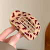 A hand holds an oval flat hair clip with a translucent pink background and dark reddish-brown leopard print pattern.