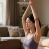 A woman is using an EMS waist pulse massager device on her abdomen while sitting on a yoga mat.