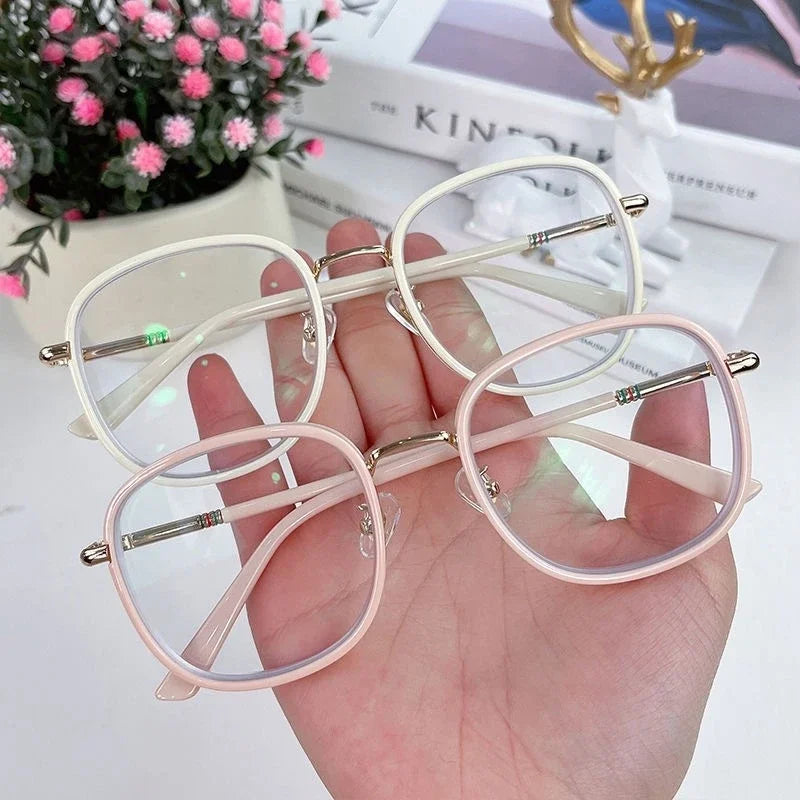 A hand holds a pair of light pink, oversized square reading glasses with silver accents.