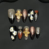 Set of 9 hand-carved press-on nails with almond petal design, gold accents, and white flowers on a black background