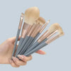 A hand holds a set of ten professional makeup brushes with light beige handles and bristles against a gray background.
