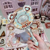 A hand holds a decorative makeup mirror set featuring a heart-shaped frame and floral accents against a pink background.