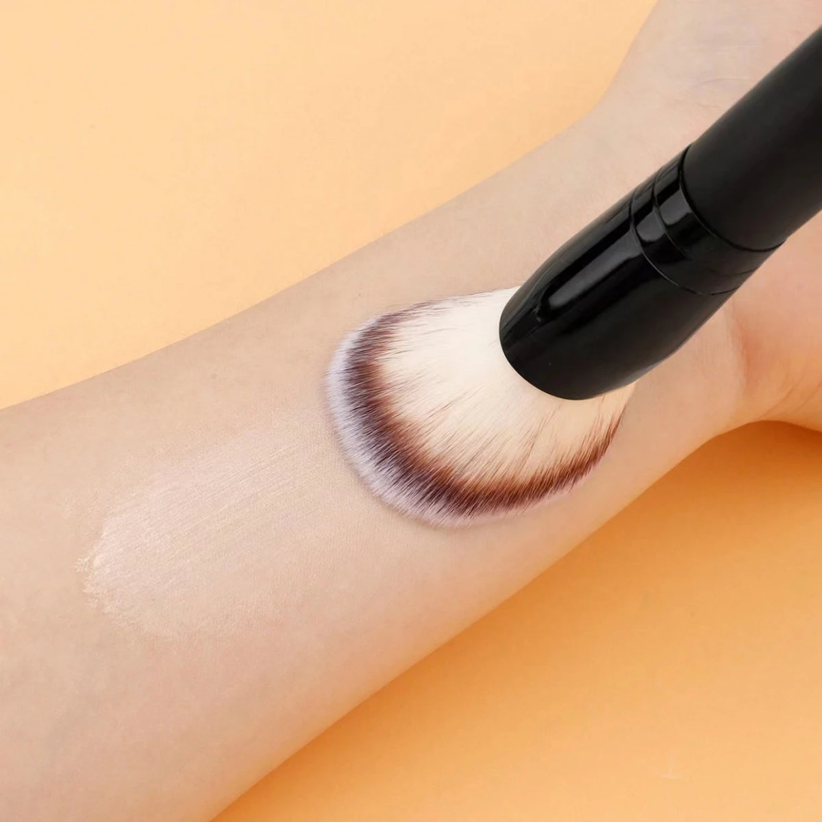 Ein Make-up-Pinsel trägt ein hellbeiges Kosmetikprodukt auf den Arm einer Person.