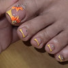 A hand displaying press-on fake nails featuring orange floral designs and gold accents on short square nails.