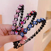 A hand holds a black rhinestone wave headband featuring pink, purple, and blue floral embellishments.