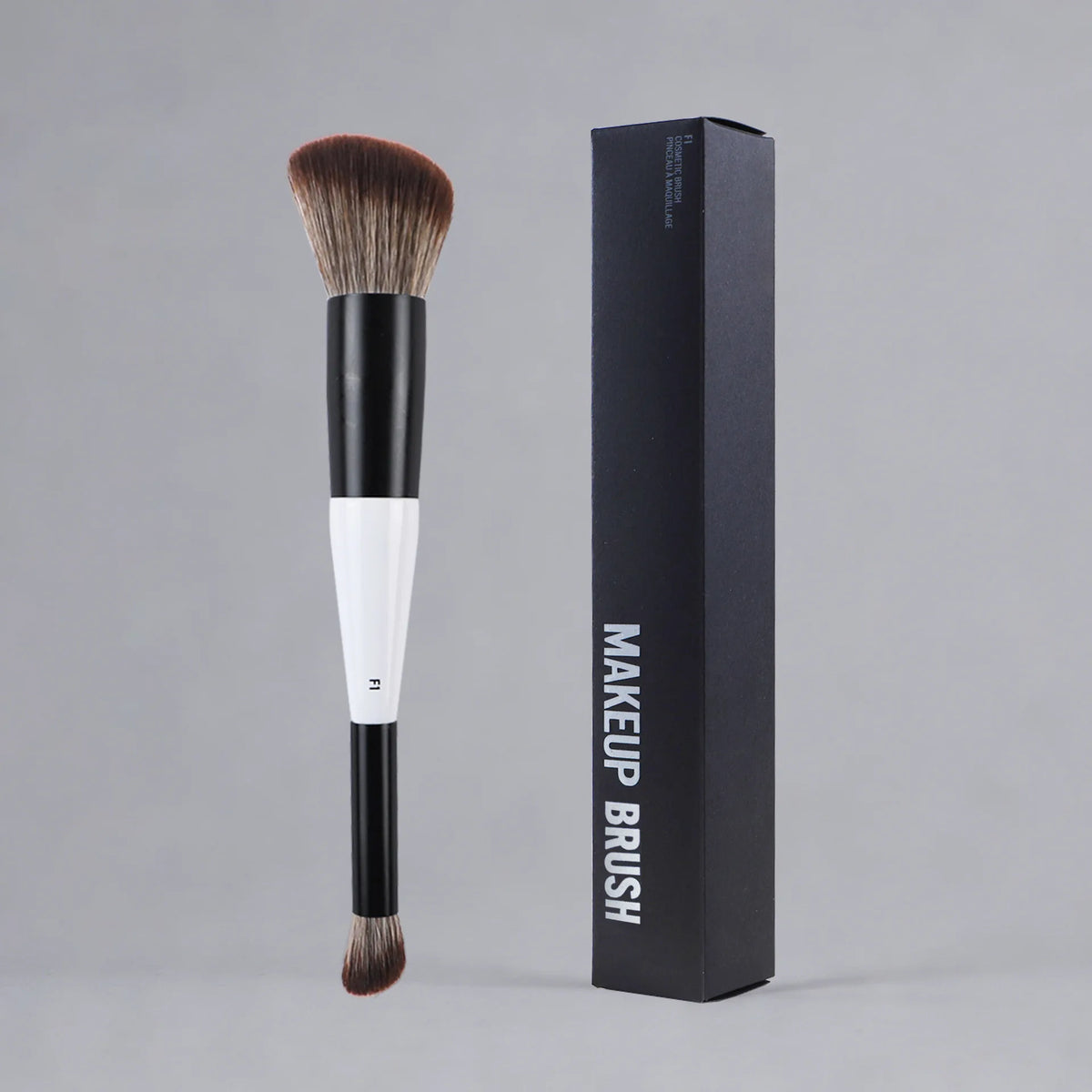 A black makeup brush with a white ferrule and bristles is shown next to its packaging box.