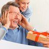 A man is using an electric hand massager on an elderly man while a younger boy watches, with a man holding a gift box.