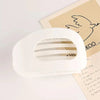 A white oval flat hair clip is shown resting on a piece of light brown paper.