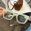 Une paire de lunettes à monture carrée rétro transparentes avec une teinte vert clair est tenue contre un arrière-plan flou.