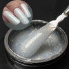 Un outil argenté plonge dans un pot de gel de construction blanc et pailleté pour extensions de ongles.