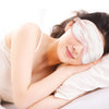A woman is relaxing with a pink silk sleep eye mask on her eyes, resting on a white pillow.