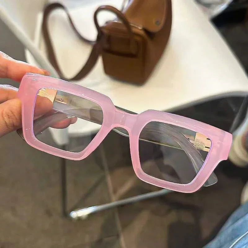 Lunettes de lecture à monture carrée rose rétro avec des verres clairs tenues par une main.