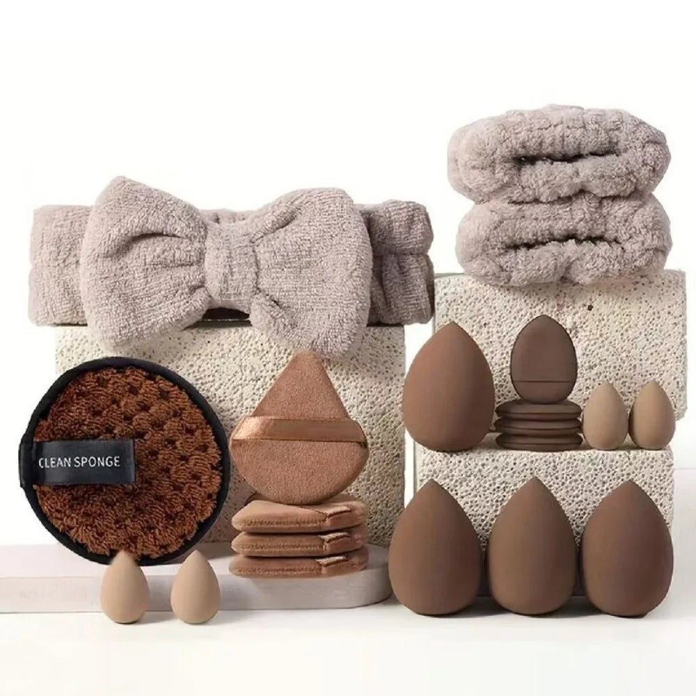 A set of makeup brushes and tools displayed on a white surface with fluffy gray fabric and brown sponges.