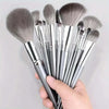 A hand holds a set of ten professional makeup brushes with silver handles and grey bristles.