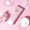 A tube of Japan Sakura Cleanser facial wash next to a glass bowl of white powder and pink flowers on a pink background.