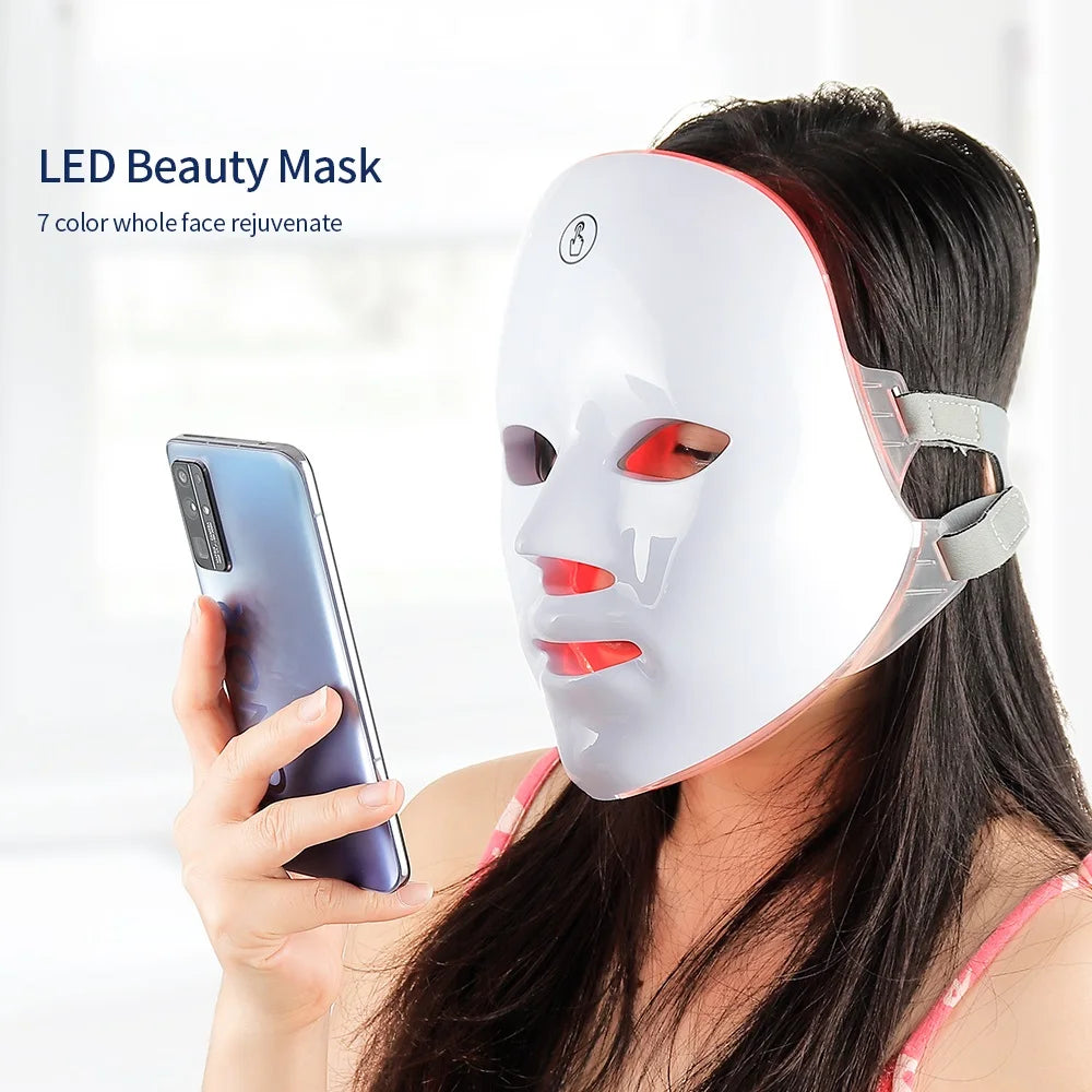 Eine Frau trägt eine weiße LED-Beautymaske auf das Gesicht und hält ein Smartphone.