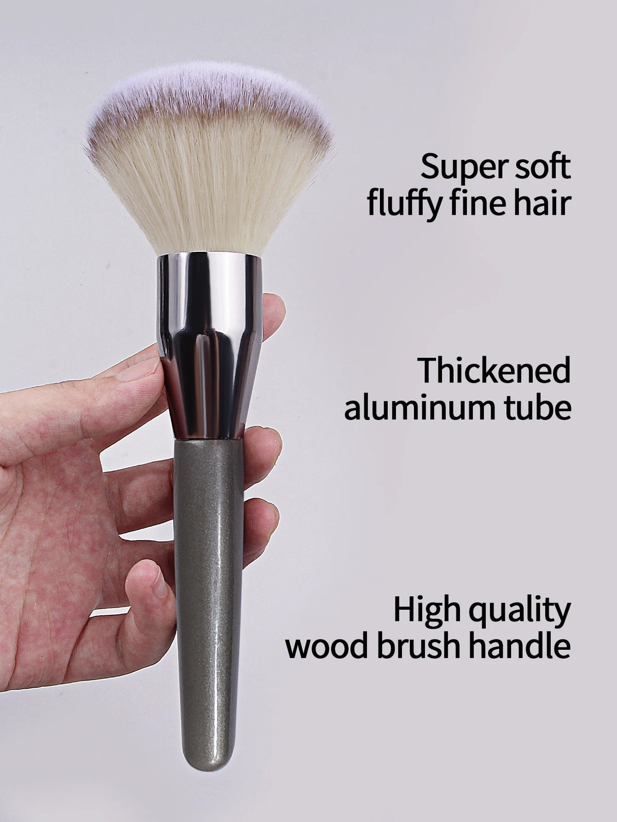 A close-up of a fluffy face powder brush with soft hair and a thickened aluminum tube, held in a hand.