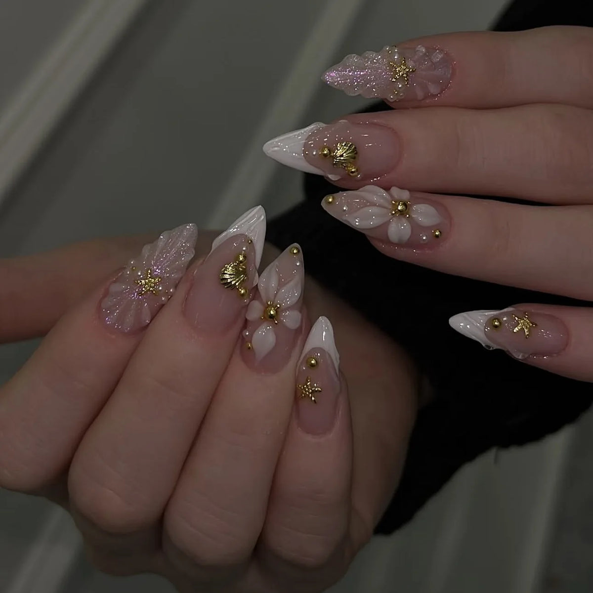 Une main montre un ensemble d'ongles press-on roses à l'amande française avec des accents floraux dorés.