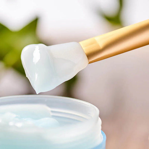 A golden silicone facial mask applicator brush is shown applying a white cream to a jar.