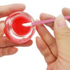 A hand is using a pink makeup brush to apply bright red liquid cosmetic onto a clear glass container.