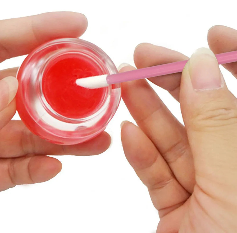 A hand is using a pink makeup brush to apply bright red liquid cosmetic onto a clear glass container.