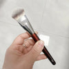Une main tient un pinceau de maquillage liquide angulé avec une ferule argentée et un manche brun sur un fond clair.