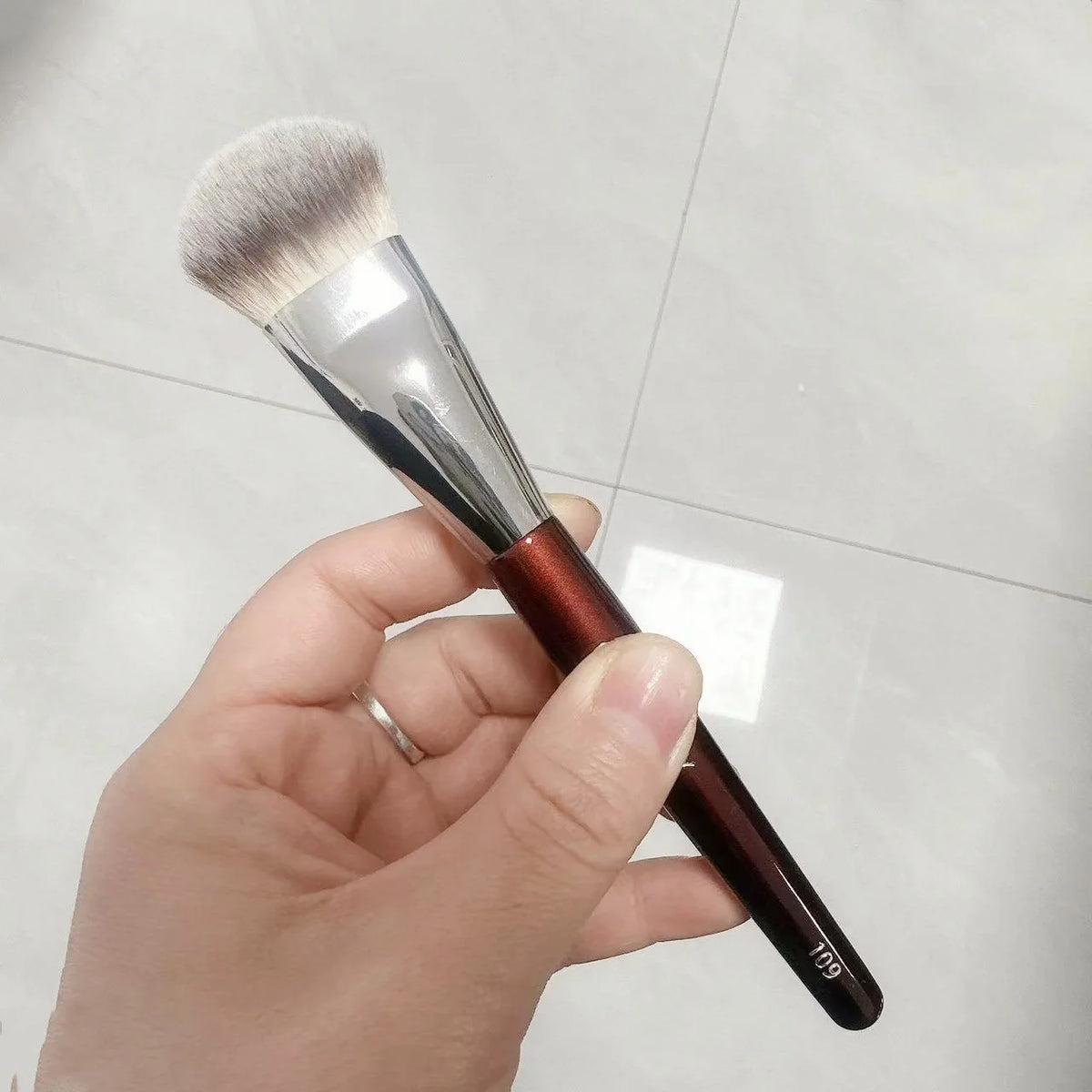 Une main tient un pinceau de maquillage liquide angulé avec une ferule argentée et un manche brun sur un fond clair.