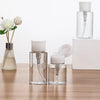 A set of clear glass refillable push-down pump dispenser bottles with white caps and a larger bottle in the background.