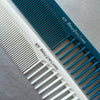 A close-up of a white and teal anti-static professional hair cutting comb with parallel teeth resting on a gray surface.