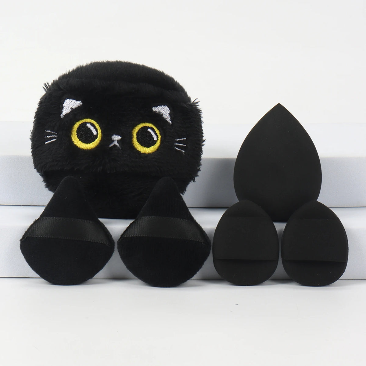 Un ensemble de poudre à tablier noire brodée, comprenant un applicateur en forme de chat et plusieurs poudres rondes.