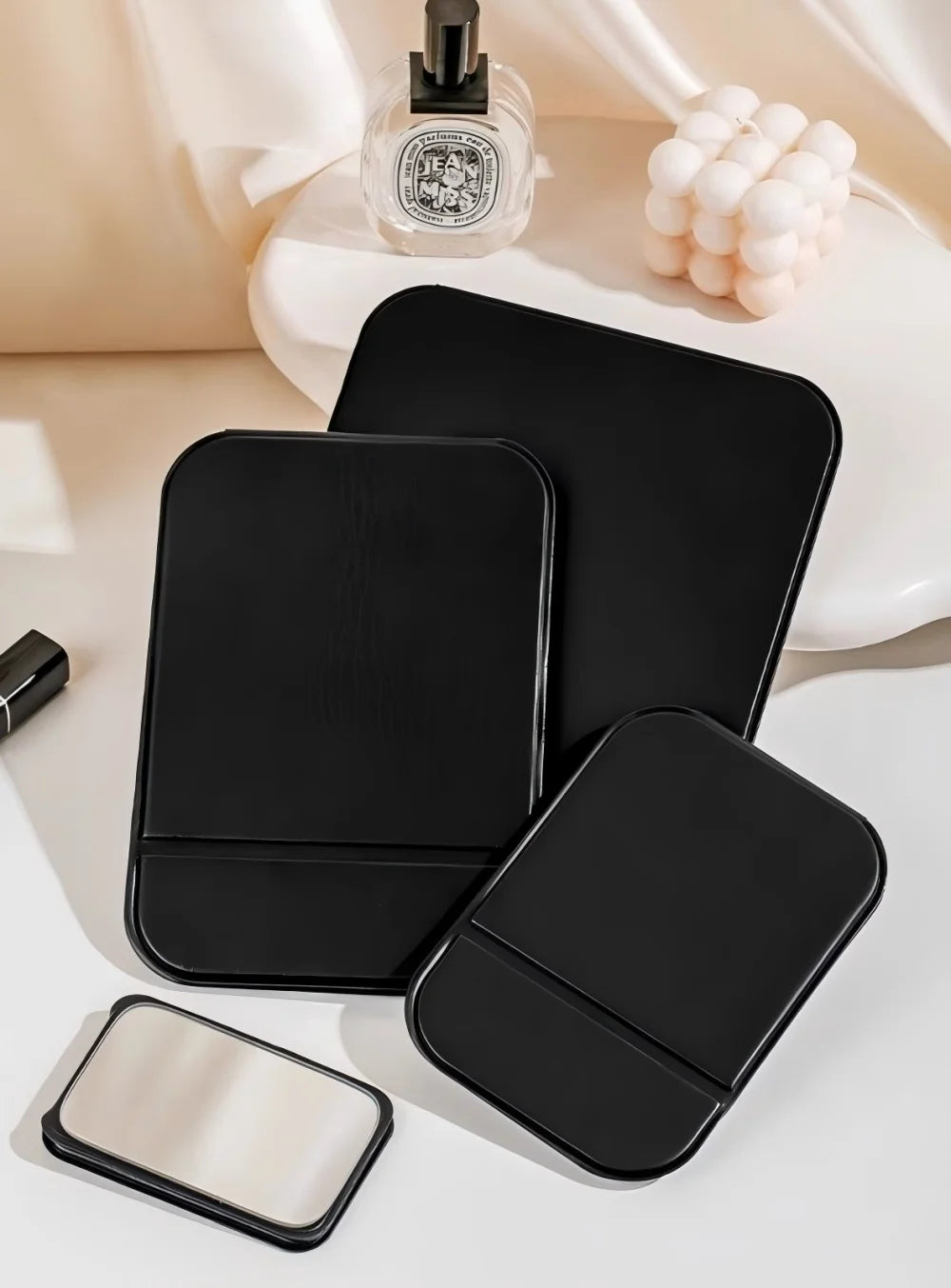 A black foldable portable makeup mirror is shown next to a round mirror and a white orb-like object.