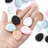 A hand holds various colored makeup sponge puffs, including black, white, pink, and beige shades.