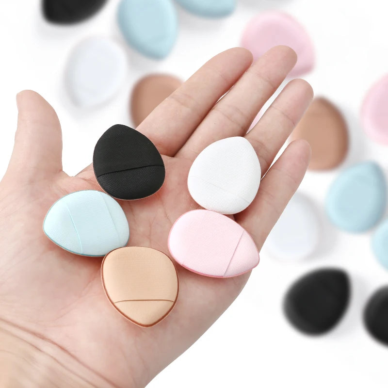 A hand holds various colored makeup sponge puffs, including black, white, pink, and beige shades.
