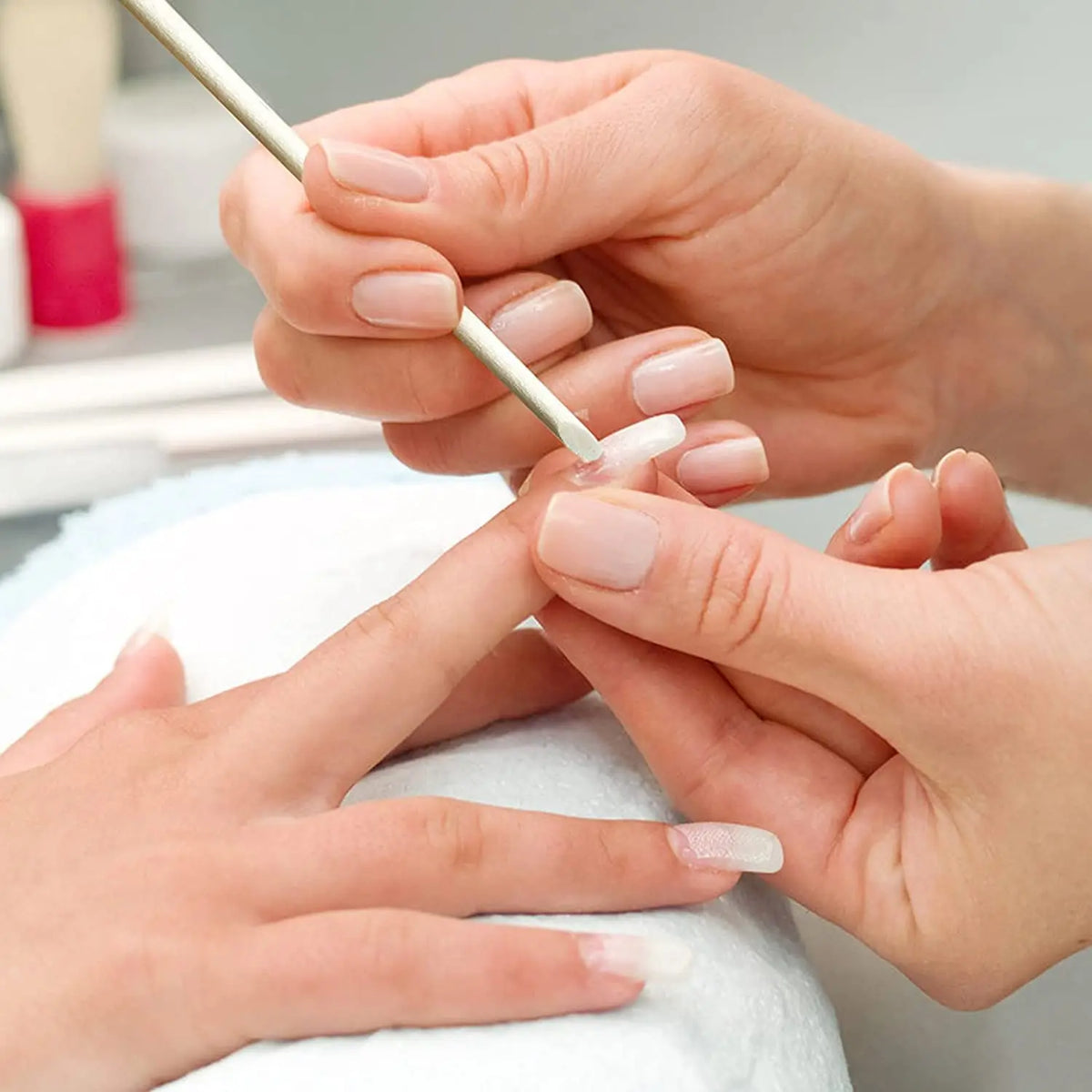 Hands are using a natural wood nail art pusher stick to apply product to a fingernail.