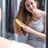 A woman is using a gold-colored scalp massage brush comb on her long brown hair.