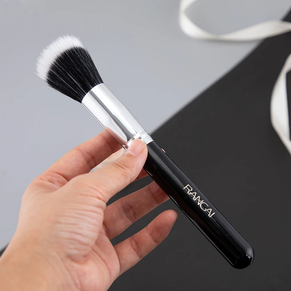 A hand holds a black and silver flat top kabuki foundation brush with white bristles against a dark background.