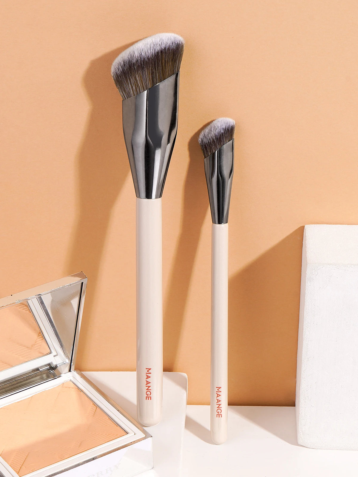 A set of two professional makeup brushes with silver ferrules and light wood handles displayed against a beige background.