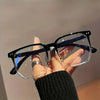 A hand holds a pair of black-framed, clear-lensed minimalist square reading glasses against a light background.
