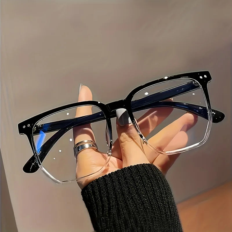 A hand holds a pair of black-framed, clear-lensed minimalist square reading glasses against a light background.