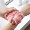 Hands are squeezing a pink exfoliating bath bubble ball with a soft mesh sponge on a white surface.