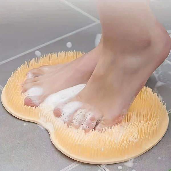 A person's foot is resting on a silicone exfoliating and massage brush, with white foam visible on the skin.