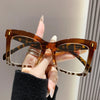 A hand holds a pair of tortoiseshell square frame reading glasses with blue light protection.