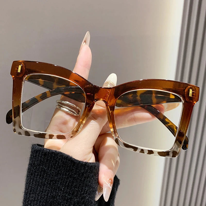 A hand holds a pair of tortoiseshell square frame reading glasses with blue light protection.