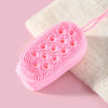 A pink silicone body brush with pink elastic attached, resting on a white towel.